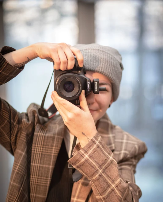 Person smiling behind a Canon DSLR camera, wearing a plaid jacket and grey beanie. Person smiling behind a Canon DSLR camera, wearing a plaid jacket and grey beanie.